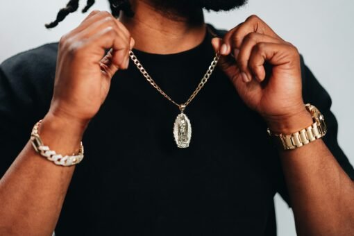Close-up of a man wearing a gold Cuban link chain with a pendant, gold watch, and bracelet against a black shirt.