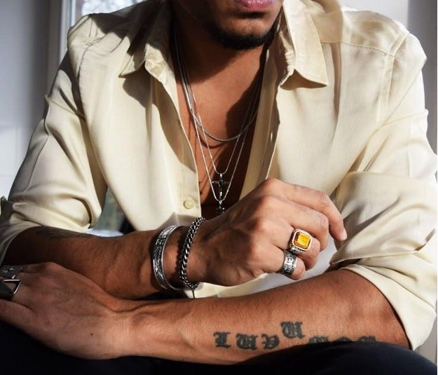 …Man wearing layered silver chains, gemstone rings, and bracelets with a light shirt and tattooed arms