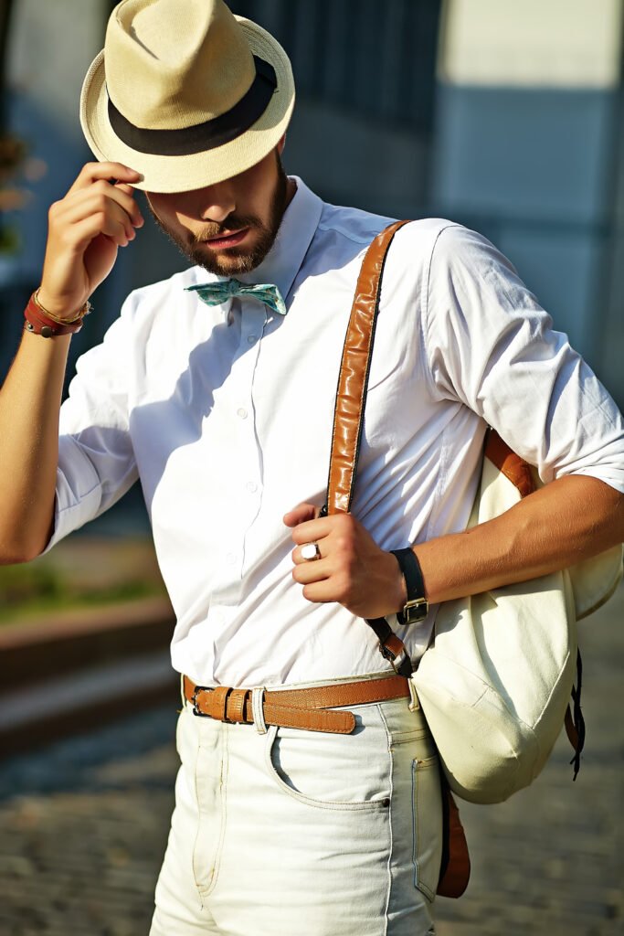 A man wearing a hat and a white shirt stands confidently, showcasing a casual yet stylish appearance